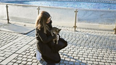 Siyah elbiseli genç bir kadın parkta bir fıskiyenin yanında kediye sarılıyor, muhtemelen hindinin içinde..