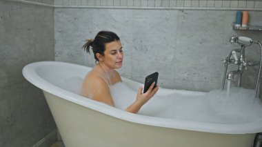 Genç bir kadın, banyo içi fayanslı baloncukların arasında akıllı telefon kullanarak bir küvette dinleniyor..
