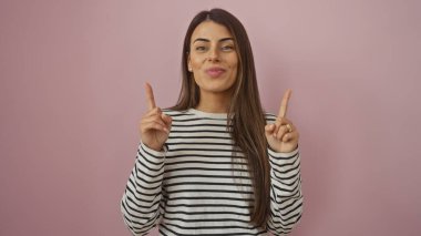 Gülümseyen genç İspanyol kadın parmaklarıyla yukarıya, pembe izole bir arka plana işaret ederek pozitifliği ve güveni temsil ediyor..