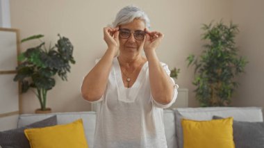 Güzel, olgun, gri saçlı, kadın, gözlük takan, ayakta, ayakta duran, içinde, oturma odası, ayarlama, çerçeveler, içinde, aydınlık, rahat, içeride, dekore edilmiş, canlı, minderlerle