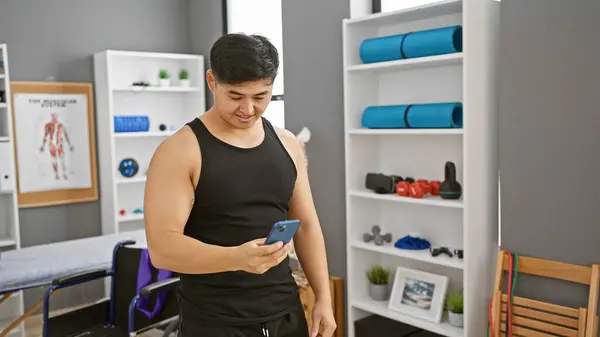 stock image Handsome asian man in a clinic interior checking his phone with fitness equipment in the background