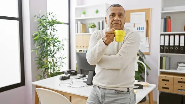 stock image A pensive middle-aged hispanic man holding a mug stands in a well-lit office with contemporary furniture and office supplies.