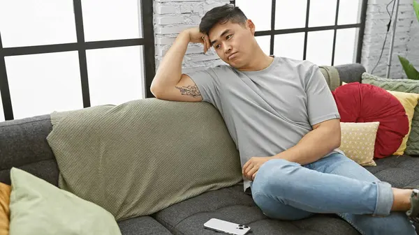 stock image Contemplative young asian man lounging on couch in a cozy, stylish apartment, embodying casual home life.