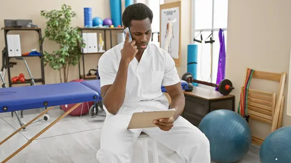 stock image A professional african man in medical attire is sitting in a rehab clinic room, examining a document and talking on the phone.