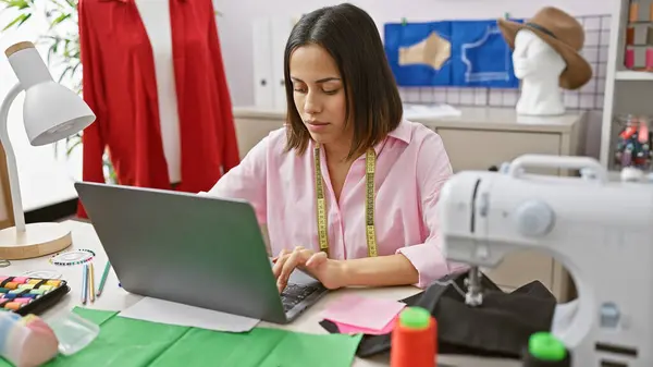 stock image A focused woman tailoring in a vibrant workshop, using a laptop next to a sewing machine on a fabric-covered table.