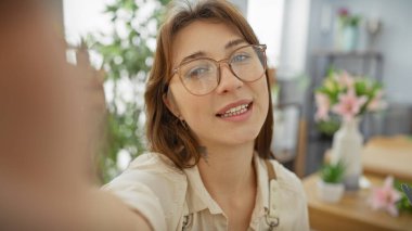 Genç beyaz bir kadın çiçekçide selfie çekiyor, güzel, kapalı, yeşil bir dükkan arka planı sergiliyor..