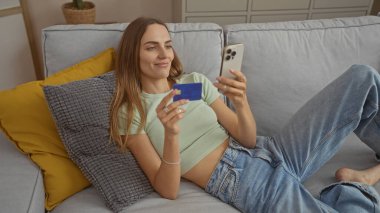Sarışın, güzel bir kadın, oturma odasındaki kanepede, elinde kredi kartı ve akıllı telefonuyla rahatlıyor..
