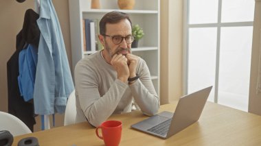 A pensive mature man with a beard working at home on his laptop in a well-lit living room. clipart