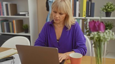 Modern bir ev ofisinde çiçekli ve kahve kupalı bir dizüstü bilgisayarda çalışan orta yaşlı bir kadın..