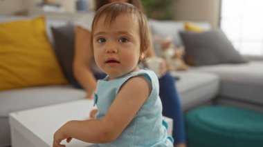 Oturma odasında arka planda bir kadınla oynayan küçük bir kız, sıcak bir aileyi sergiliyor, evde sevgi ve beraberlikle oturuyor..
