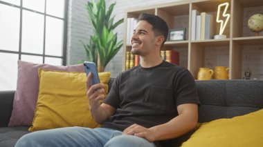 Modern bir oturma odasında akıllı telefon kullanan sakallı, genç İspanyol bir adam..