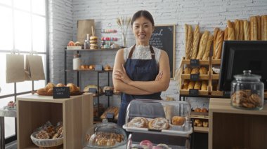 Çeşitli ekmek ve hamur işleriyle fırındaki genç Çinli kadın, Çin 'de kolları çaprazlanmış vaziyette, iç mekanda önlük takıyor.