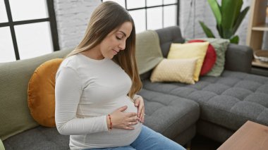 Sakin bir İspanyol kadın, hamile, modern bir oturma odasında oturuyor, sıcaklığı ve anneliği çağrıştırıyor..