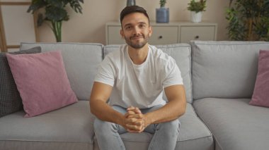 Evinin oturma odasında sakallı yakışıklı, beyaz tişört ve kot pantolon giyerken rahat ve neşeli görünen genç bir İspanyol..