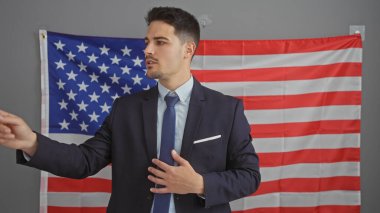 Takım elbiseli yakışıklı genç adam, Amerikan bayrağı arkaplanı olan bir ofiste özgüvenle el kol hareketi yapıyor.