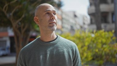 Sıradan giysili yakışıklı kel adam yemyeşil bir şehir bahçesinde dikilirken dalgın dalgın bakıyor..