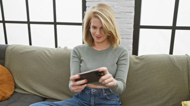 Beyaz kadın, kısa sarı saçlı. Kanepede akıllı telefon kullanıyor.