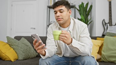 Modern bir oturma odasında kahve ve akıllı telefonla rahatlayan genç bir adam..