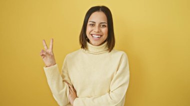 Sarı arka planda rahat süveter giyen mutlu, genç Latin bir kadın. Parlak bir gülümsemeyle barış işareti gösteriyor.