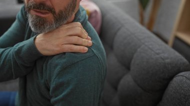Sıradan giyinmiş sakallı bir adam evinde koltukta omuz ağrısı çekiyor..