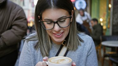 Gülümseyen genç bir kadın kalabalık bir terasta bir fincan kahvenin tadını çıkarıyor..