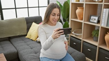 Evde oturan gülümseyen hamile bir kadın modern bir oturma odasında akıllı telefon kullanıyor..