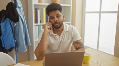 Telefondaki yakışıklı siyah adam evde dizüstü bilgisayar kullanıyor, dövmeli, odaklanmış ve ciddi..