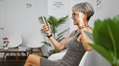 A cheerful mature woman with grey hair using a smartphone in a bright indoor waiting room setting. clipart