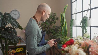 Sakallı kel adam çeşitli bitkilerle dolu bir çiçekçide akıllı telefon kullanıyor.