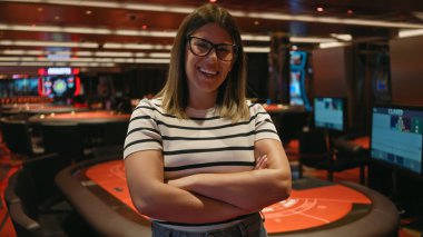 Gözlüklü, gülümseyen, genç, esmer bir kadın, hareketli bir kumarhane ortamında güvenle ayakta duruyor..
