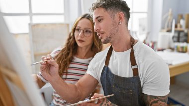 Gündelik giysiler içinde bir kadın ve bir erkek birlikte parlak bir sanat stüdyosunda resim yapıyorlar..