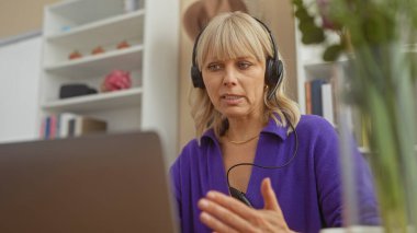 Sarışın, beyaz bir kadın evde bir video görüşmesi yapıyor. Oturma odasında kulaklık takıyor.