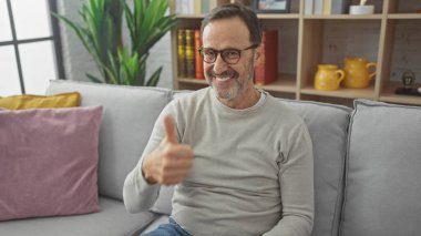 Gözlüklü, gülümseyen, olgun bir adam, rahat bir oturma odasında gri bir kanepede otururken baş parmağını kaldırıyor..