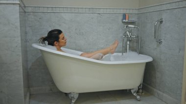 Rahat bir kadın mermer bir banyoda eski bir pençe küvetinde köpük banyosunun keyfini çıkarıyor..