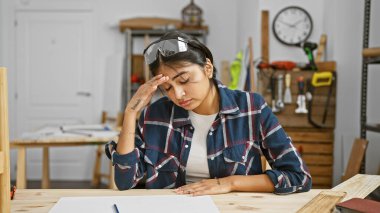 A thoughtful south asian woman in a workshop with protective glasses resting on her head contemplates her carpentry work. clipart