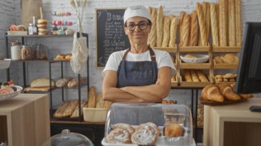 Ekmek, hamur işi ve kurabiyelerle dolu bir fırında kolları çapraz duran, gözlüklü, beyaz şapkalı orta yaşlı bir kadın, iş ortamında profesyonel ve kendinden emin bir görünüm sergiliyor..