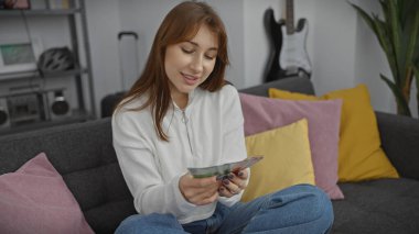 Genç beyaz kadın, renkli yastıklarla çevrili bir kanepede otururken Norveç kronunu sayıyor..