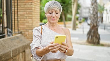 Kısa gri saçlı ve güzel gülüşlü olgun bir İspanyol kadın güneşli bir şehir sokağında dururken akıllı telefonuna odaklanıyor..