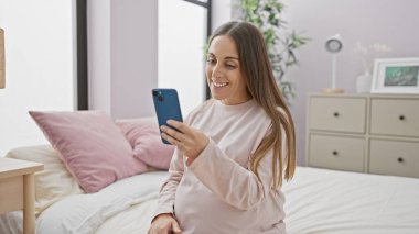 Latin kökenli hamile bir kadın yatak odasında akıllı telefon kullanırken gülümsüyor. Evde rahat bir annelik hayatı sunuyor..