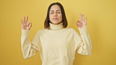Süveter içinde, sarı bir arka plana karşı meditasyon yapan, sükunet ve farkındalığı somutlaştıran sakin bir İspanyol kadın..