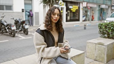 Şehirde bir bankta oturan genç bir kadın, arka planda dükkanları ve motosikletleri olan telefonunu kullanıyor..