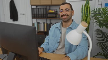 Ofis ortamında çiçekler, kitaplıklar ve masa lambasıyla gülümseyen gözlüklü İspanyol bıyıklı adam..