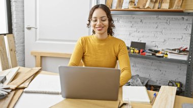 Marangozluk atölyesinde dizüstü bilgisayarı, aletleri ve odunları olan gülümseyen bir girişimci kadın..