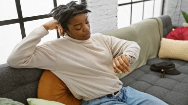 Rasta bukleli genç bir kadın, oturma odasındaki kanepede dinlenirken zamanı kontrol ediyor..