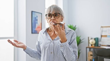 Mature hispanic woman with grey hair sending a voice message in an office interior. clipart