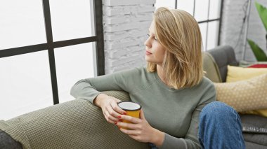 Sarışın bir kadın elinde kahve fincanıyla evinde dinleniyor.