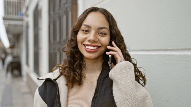 Genç, gülümseyen, İspanyol bir kadın, şehir caddesinde dururken cep telefonuyla konuşuyor..