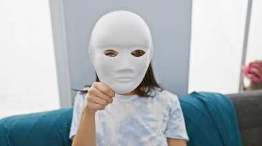 A young woman holds a blank white mask in a modern living room, suggesting anonymity or mystery. clipart