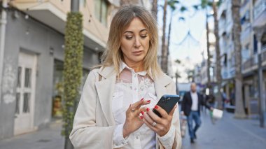 Kafkasyalı sarışın kadın güneşli bir şehirde akıllı telefon kullanıyor.