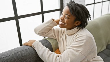 Rastalı neşeli bir Afrikalı kadın, kazak giyiyor, rahat bir oturma odasındaki kanepede rahatlıyor, mutluluk ve rahatlık saçıyor..